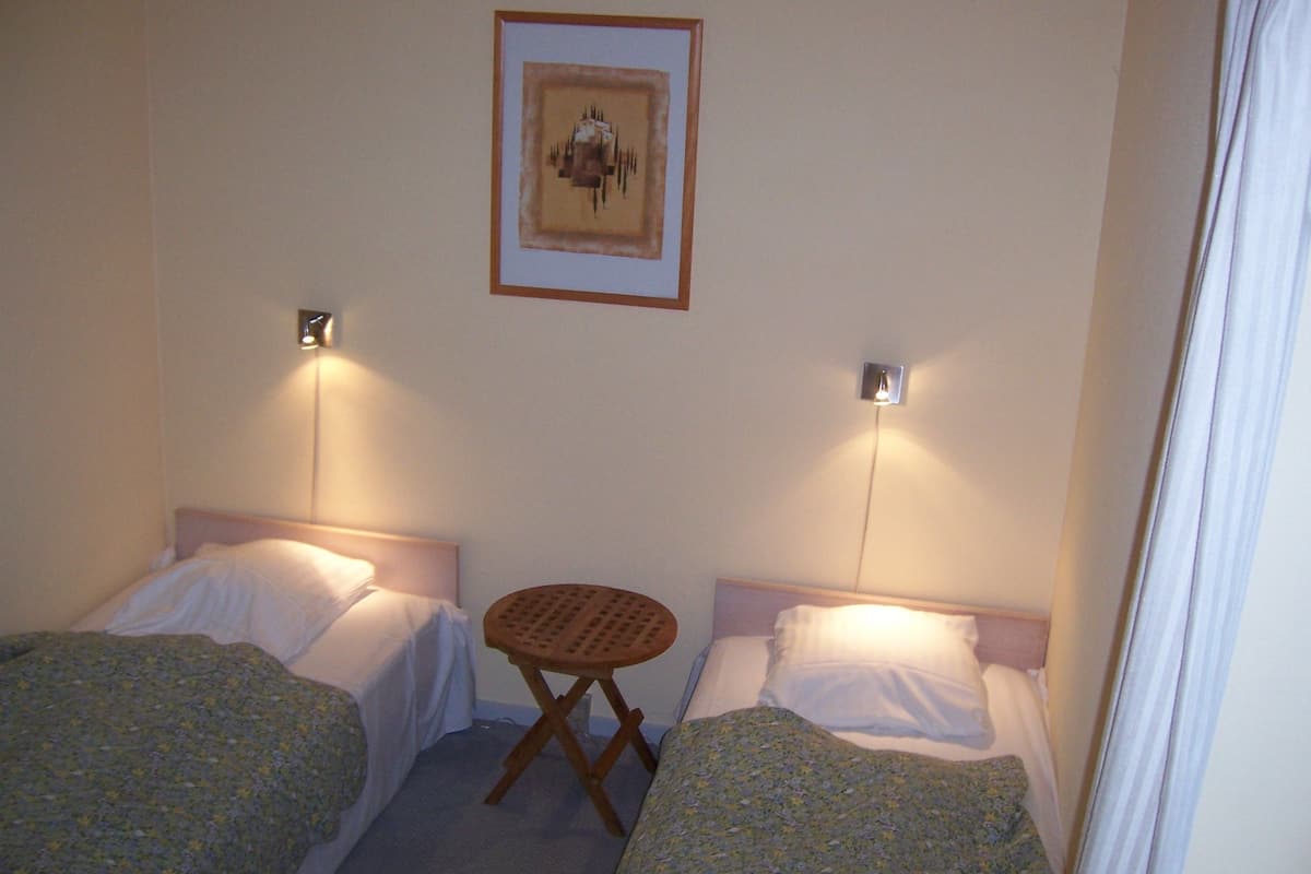 Um quarto pequeno e pouco iluminado, com duas camas de solteiro, uma pequena mesa de madeira e um quadro na parede.