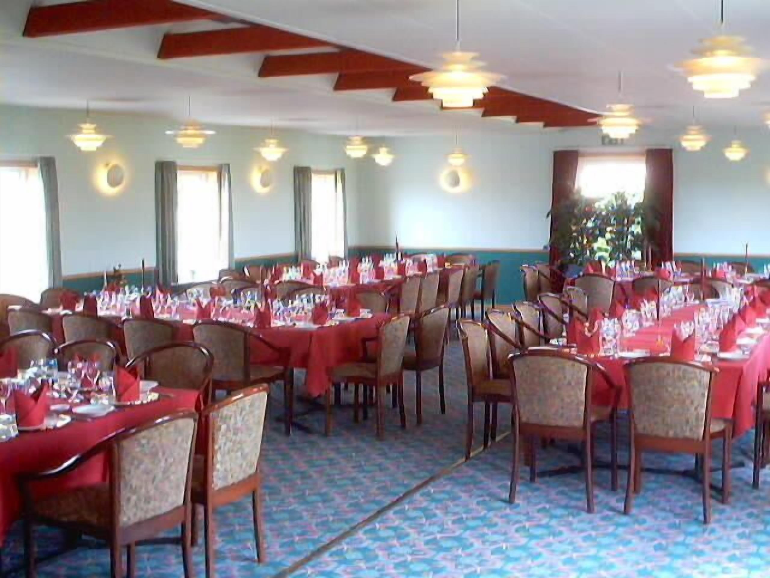 Een balzaal met gedekte tafels voor een formeel evenement, met houten stoelen en rode tafelkleden.