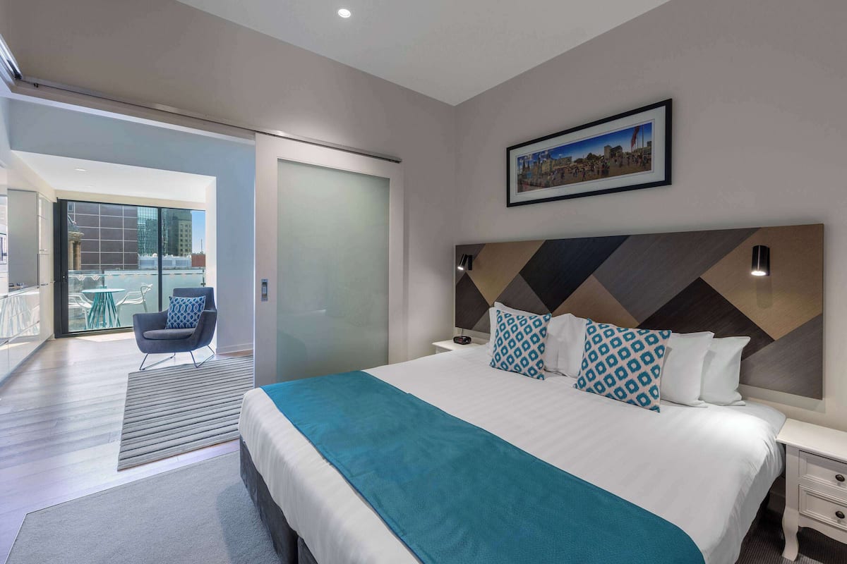 A modern hotel room with a large bed, a view of the city, and a balcony with a chair.
