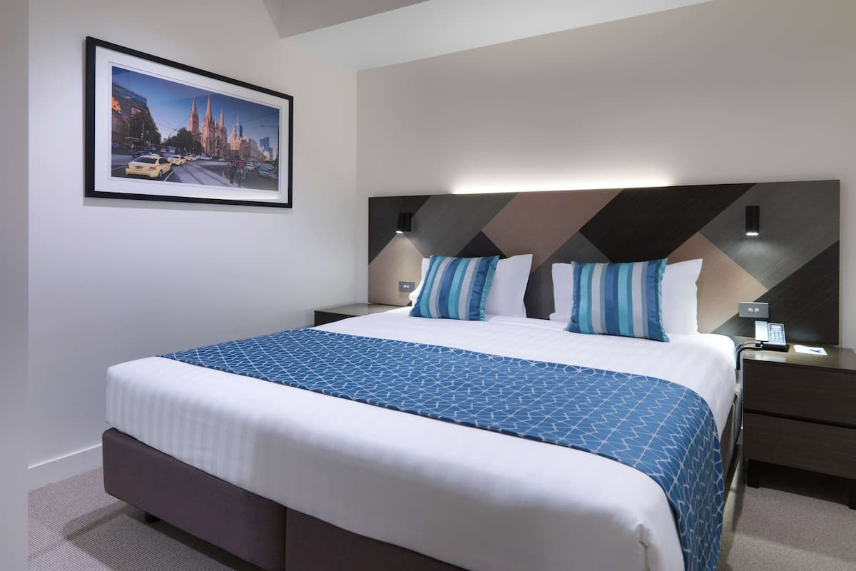 A hotel room with a large bed, a headboard with geometric patterns, a nightstand with a lamp, and a framed picture of a cityscape on the wall.