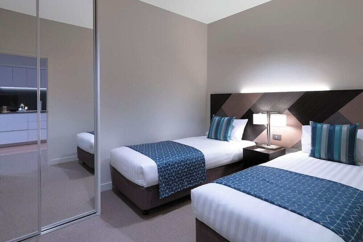Una habitación de hotel moderna con dos camas, un separador de vidrio y una kitchenette al fondo.