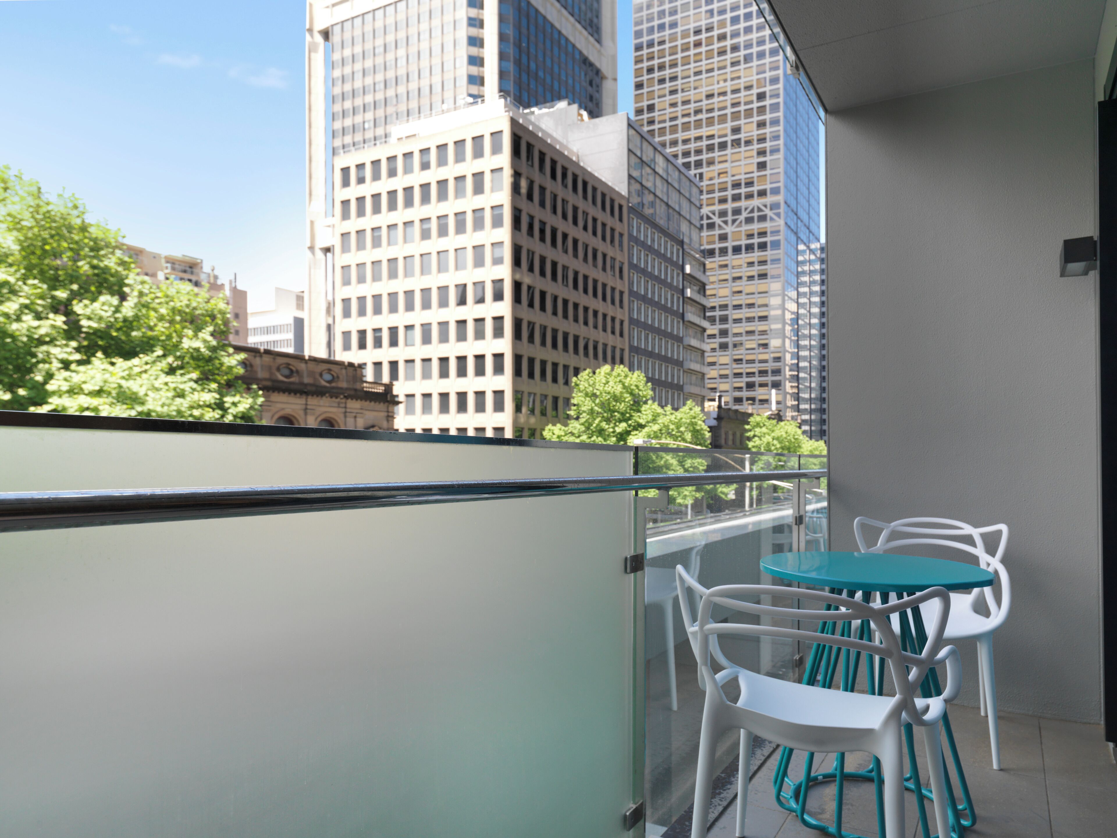 Un balcon doté d’une balustrade en verre, de chaises blanches et d’une table de couleur turquoise. En arrière-plan, on aperçoit de grands immeubles et des arbres.