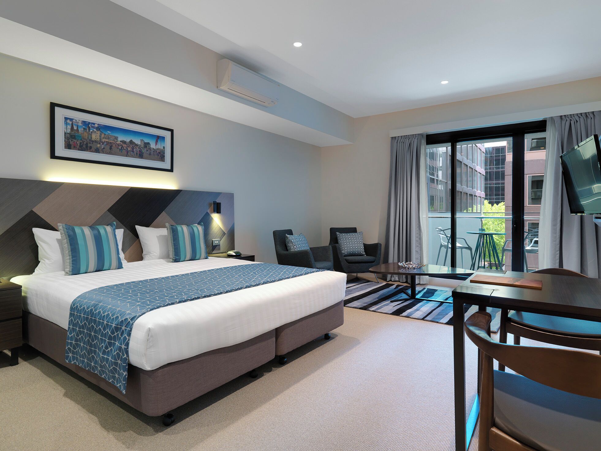 A hotel room with a bed, a desk, a chair, a TV, and a balcony with a view of buildings.