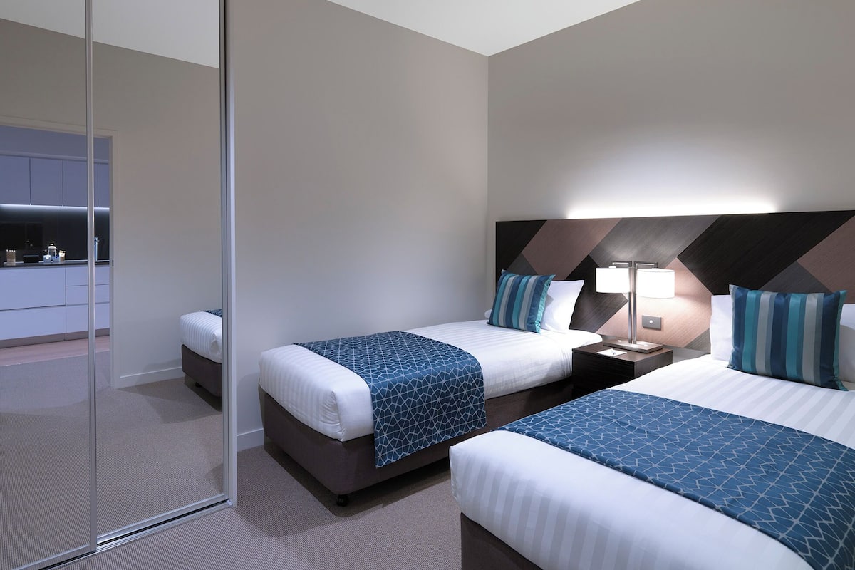A modern hotel room with two beds, a glass partition, and a kitchenette in the background.