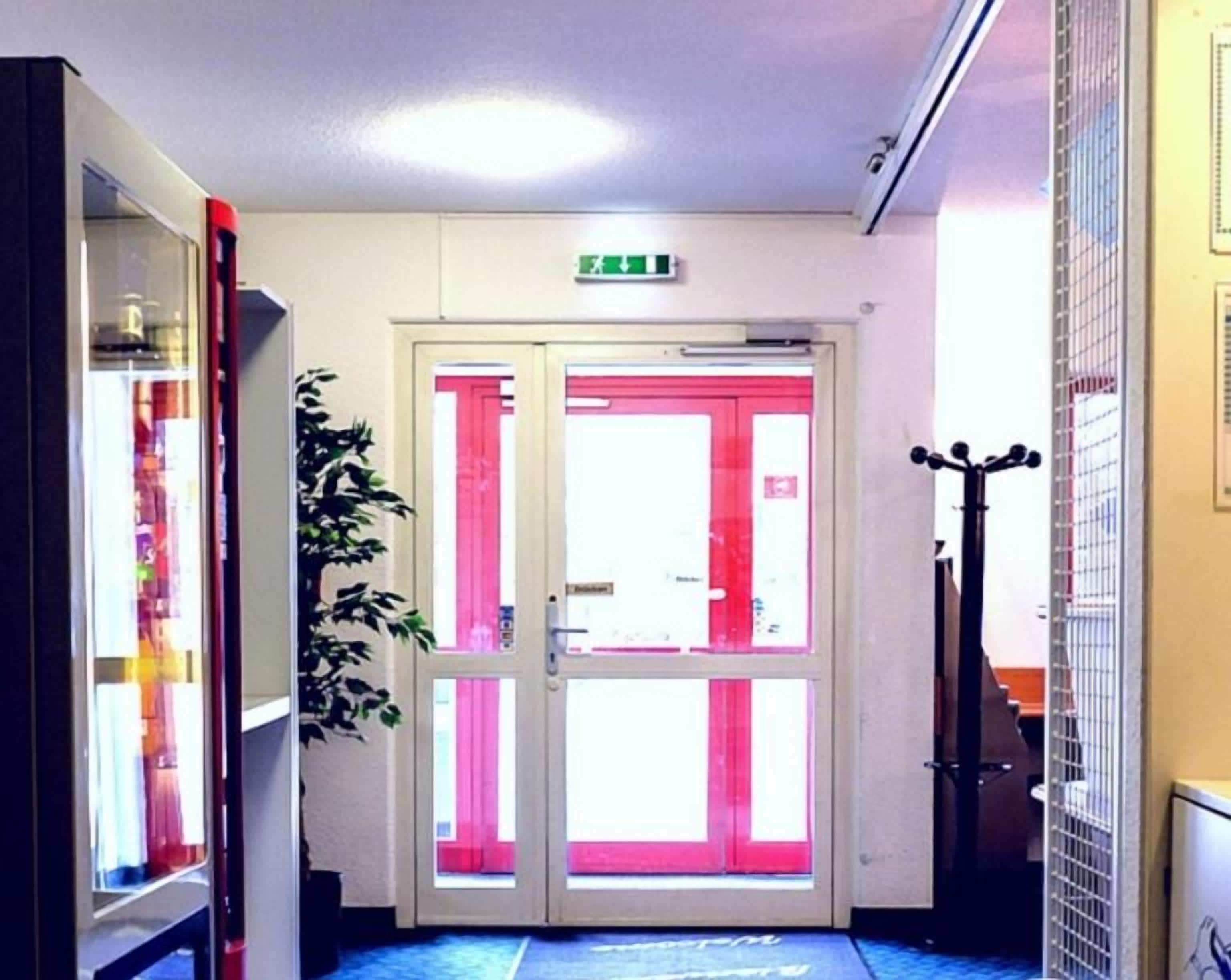 Un pasillo con piso azul estampado, una puerta doble con detalles rosados y un cartel que indica motel las 24 horas.