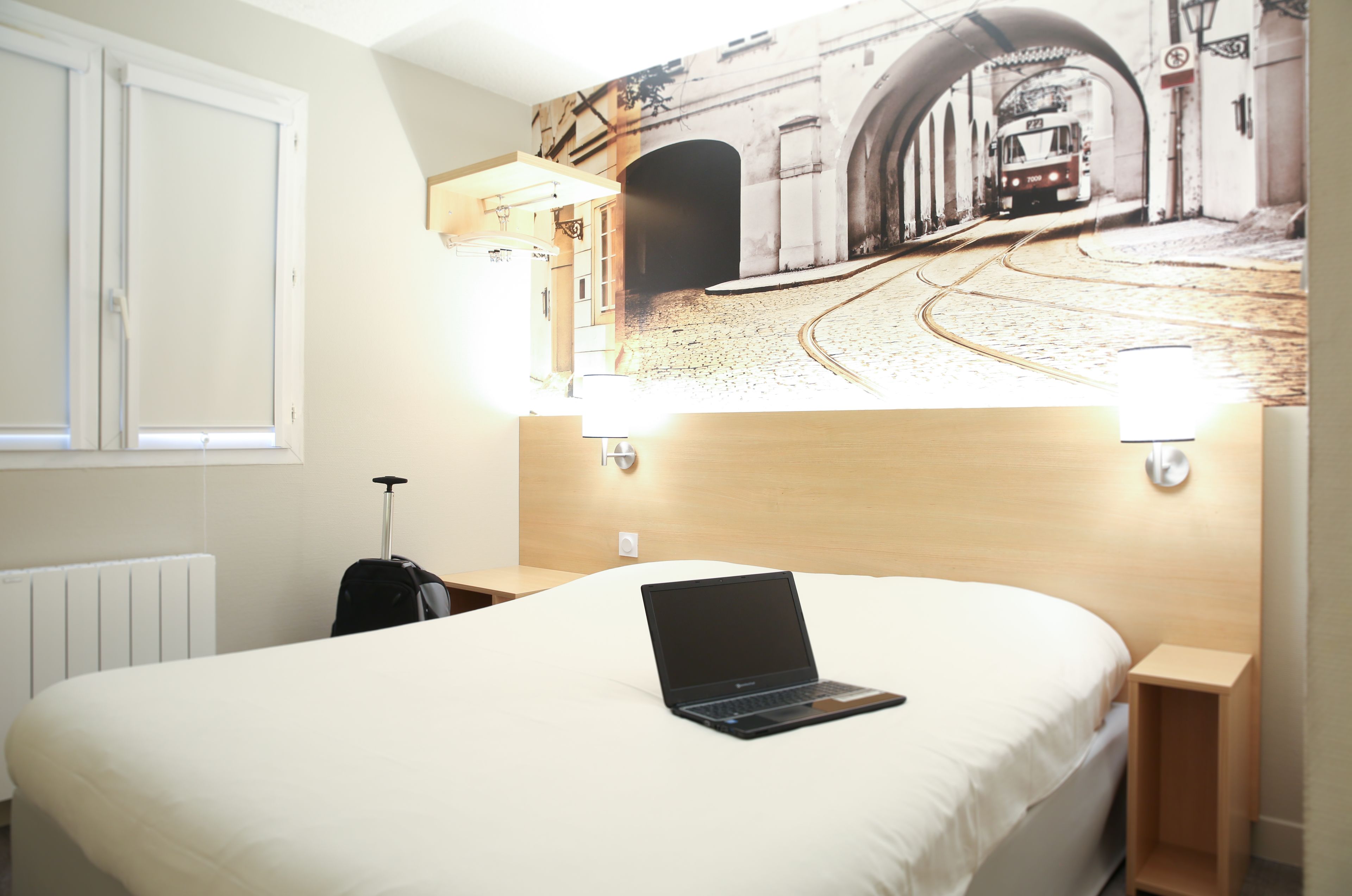 A hotel room with a bed, a laptop on the bed, a suitcase, and a wall mural of a street scene.