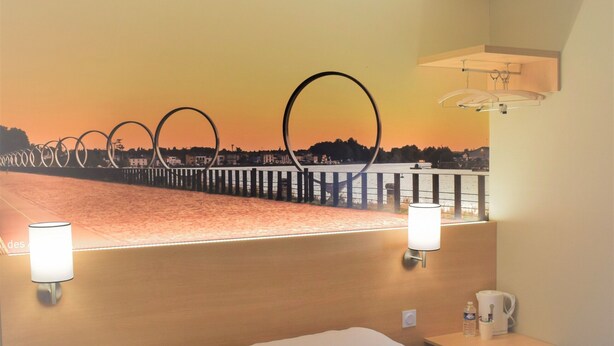 A hotel room with a bed, two pillows, a nightstand, a lamp, and a view of a sunset over a body of water. - Hôtel Inn Design Châteaubriant (Chateaubriant)