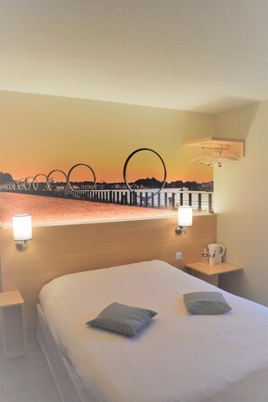 A hotel room with a bed, two pillows, a nightstand, a lamp, and a view of a sunset over a body of water. - Hôtel Inn Design Châteaubriant (Chateaubriant)