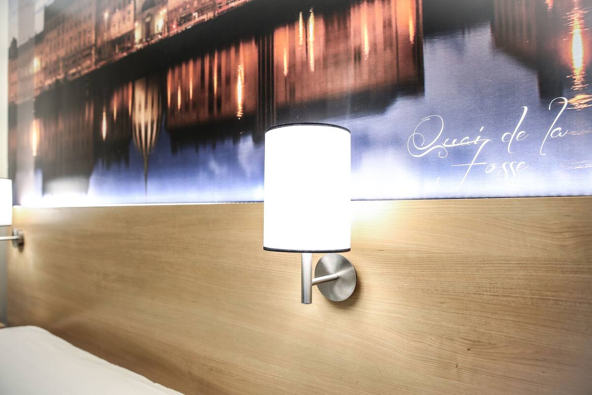 A modern hotel room with a wooden headboard, a wall-mounted lamp, and a large mural depicting a cityscape with the text "Que sais-je de la Fosse".