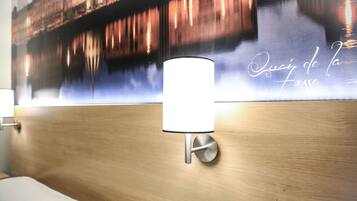 A modern hotel room with a wooden headboard, a wall-mounted lamp, and a large mural depicting a cityscape with the text "Que sais-je de la Fosse".
