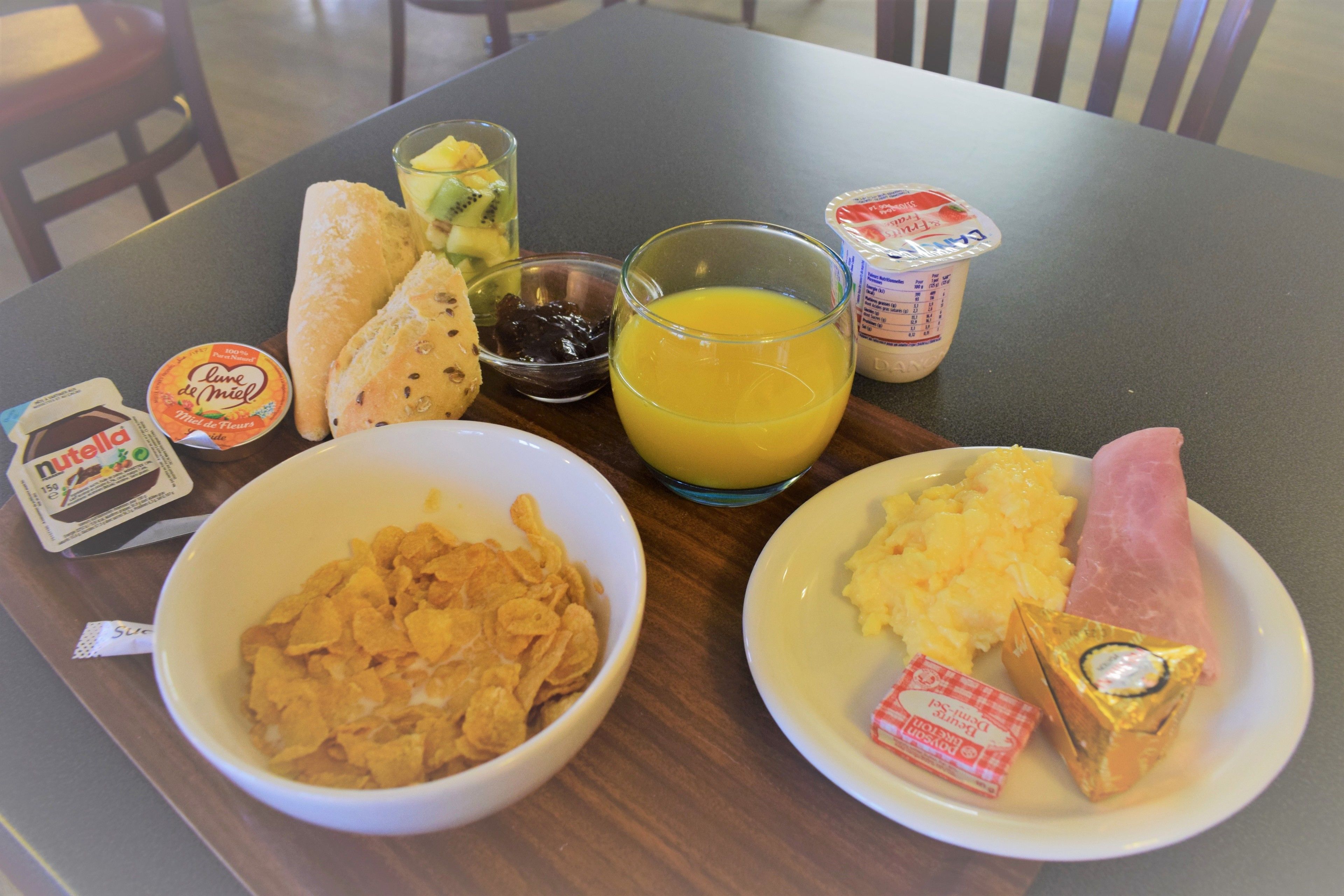 Bandeja de desayuno con cereal, tostadas, fruta, jugo, yogur y un sándwich de jamón.