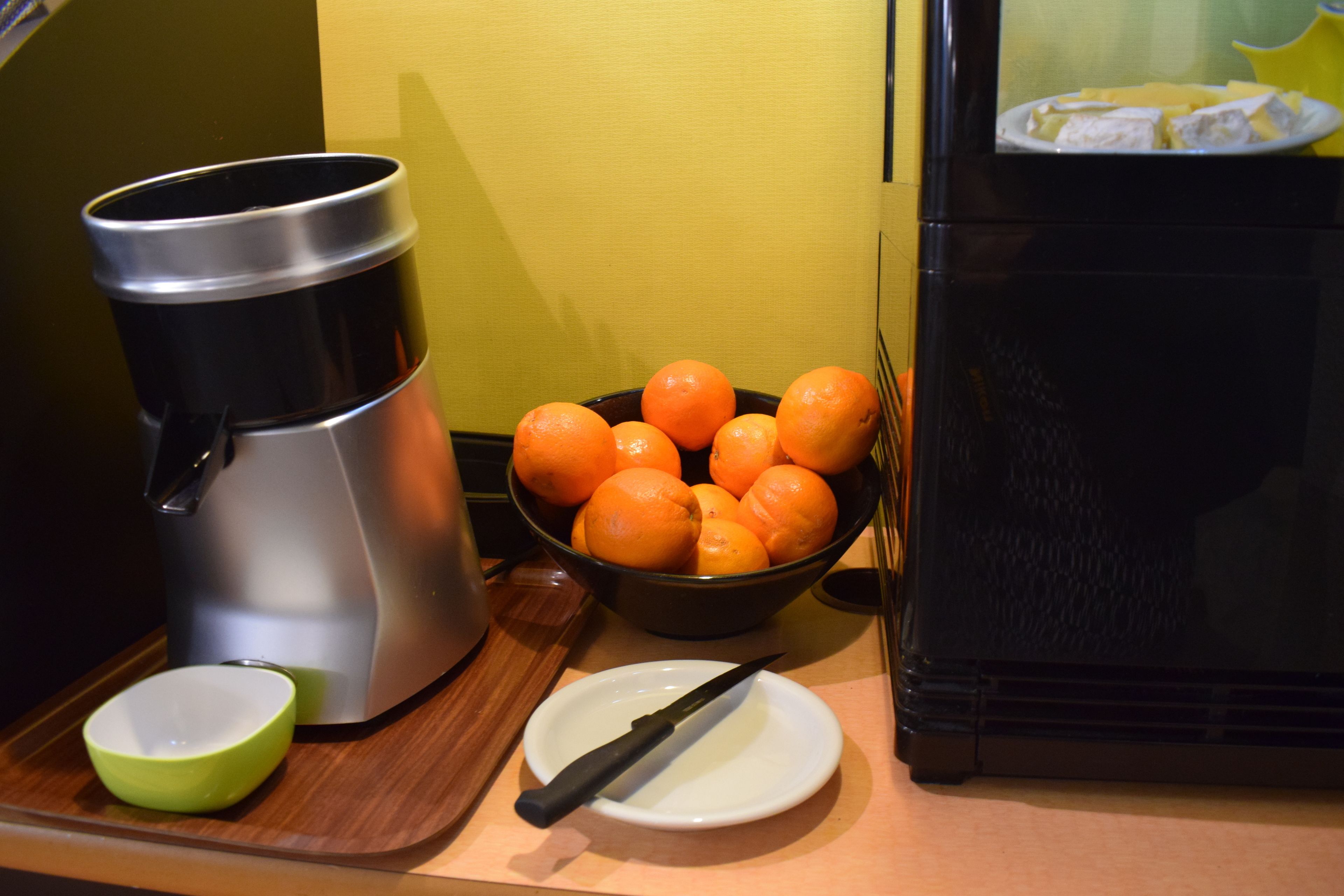 Un comptoir de cuisine avec une machine à café, un bol d’oranges, un bol vert et une assiette blanche avec un couteau.