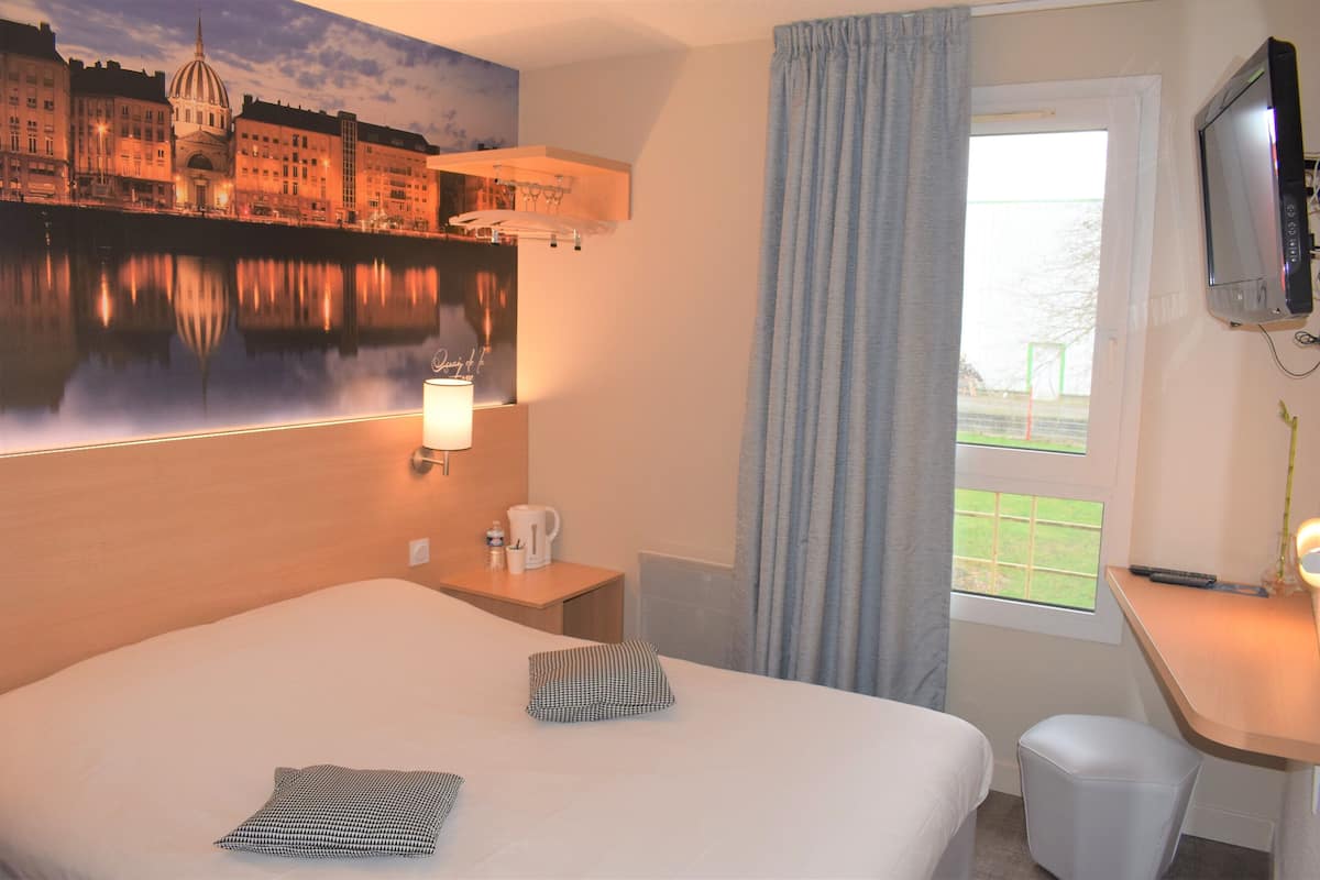 A hotel room with a bed, a desk, a TV, a window with curtains, and a painting of a cityscape.