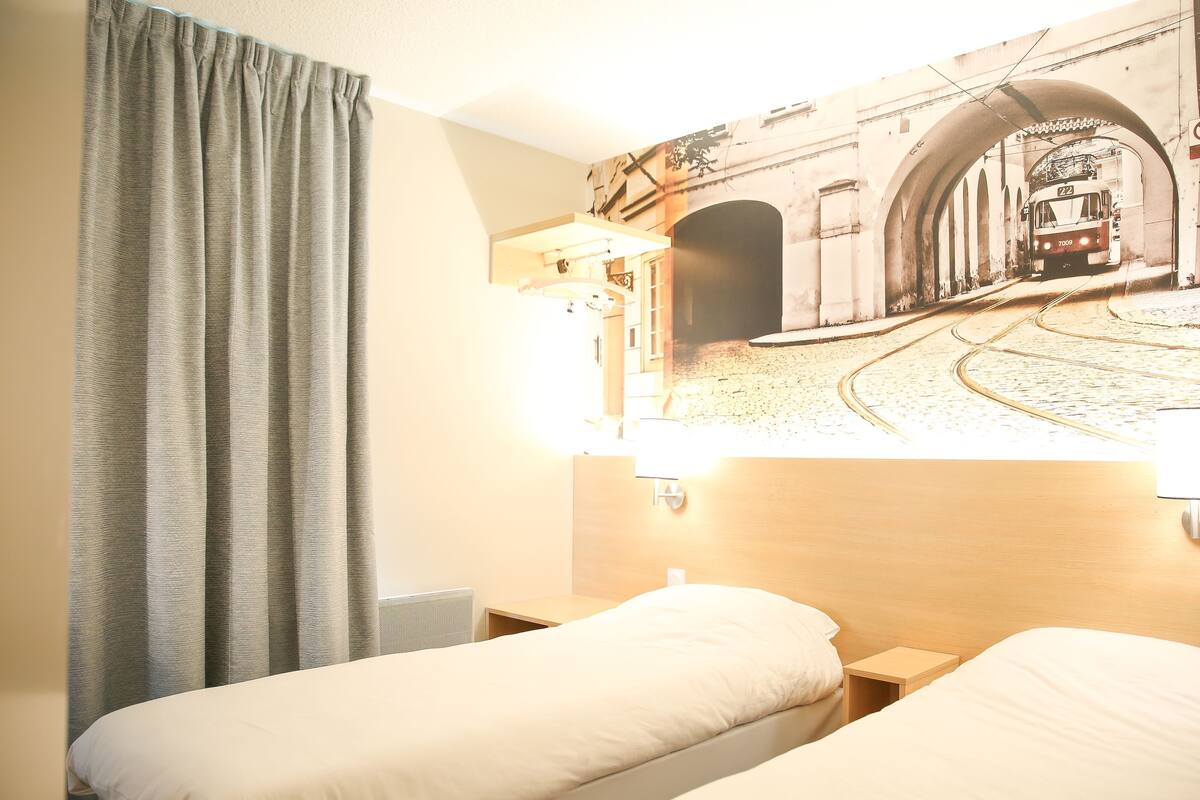 A hotel room with two beds, a headboard with a cityscape mural, a window with curtains, and a nightstand with a lamp.