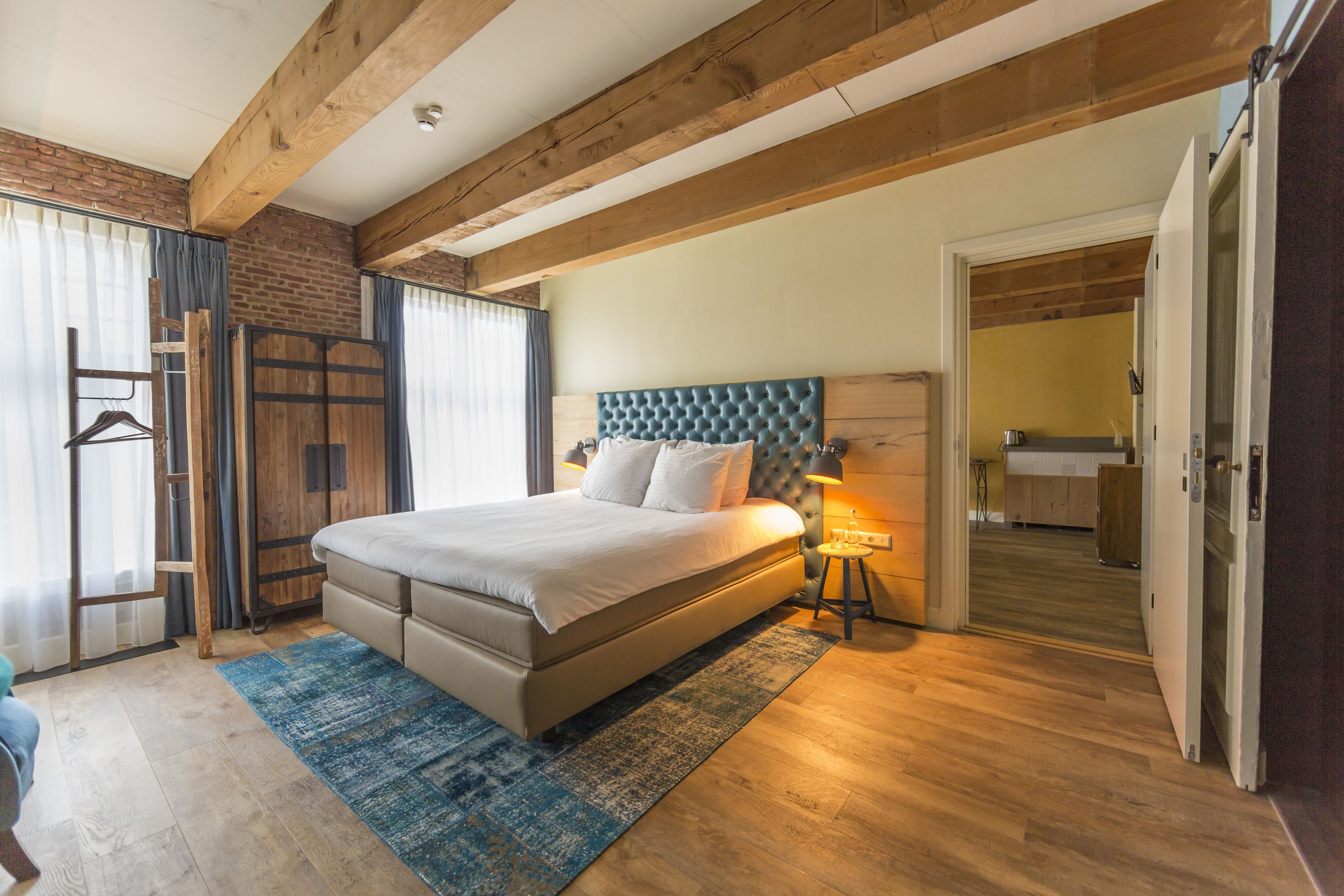 A bedroom with a large bed, wooden beams, a brick wall, and a view into the kitchen.