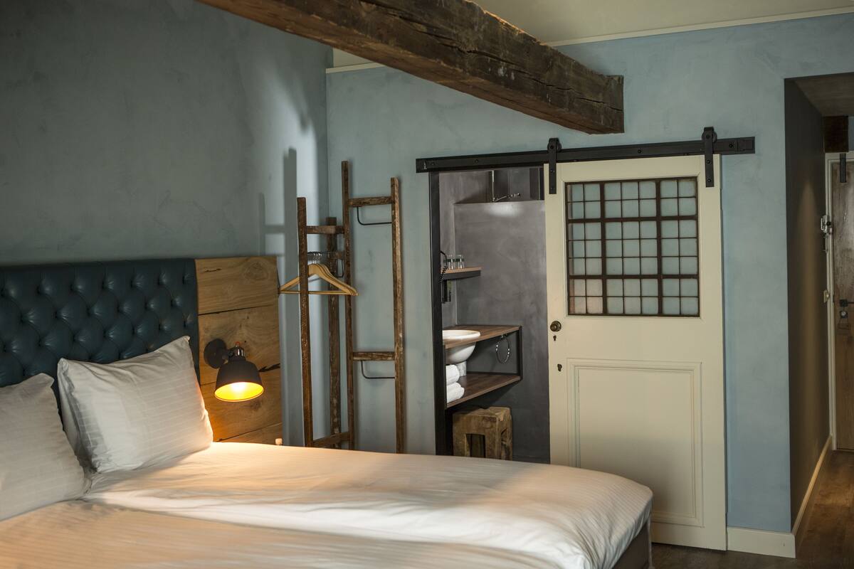 A bedroom with a large bed, a wooden headboard, a hanging light fixture, a sliding door, and a small wooden stool.