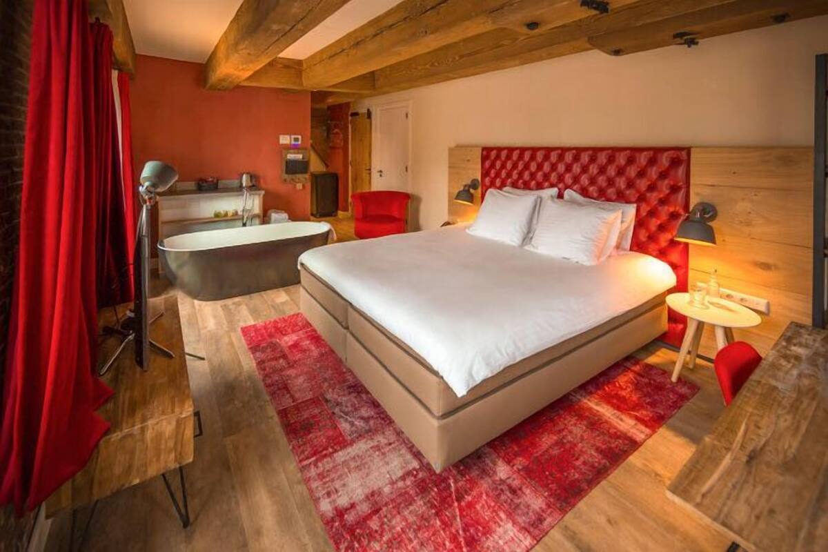 A bedroom with a large bed, a red headboard, a red rug, a wooden floor, a red chair, a small table, and a red curtain.