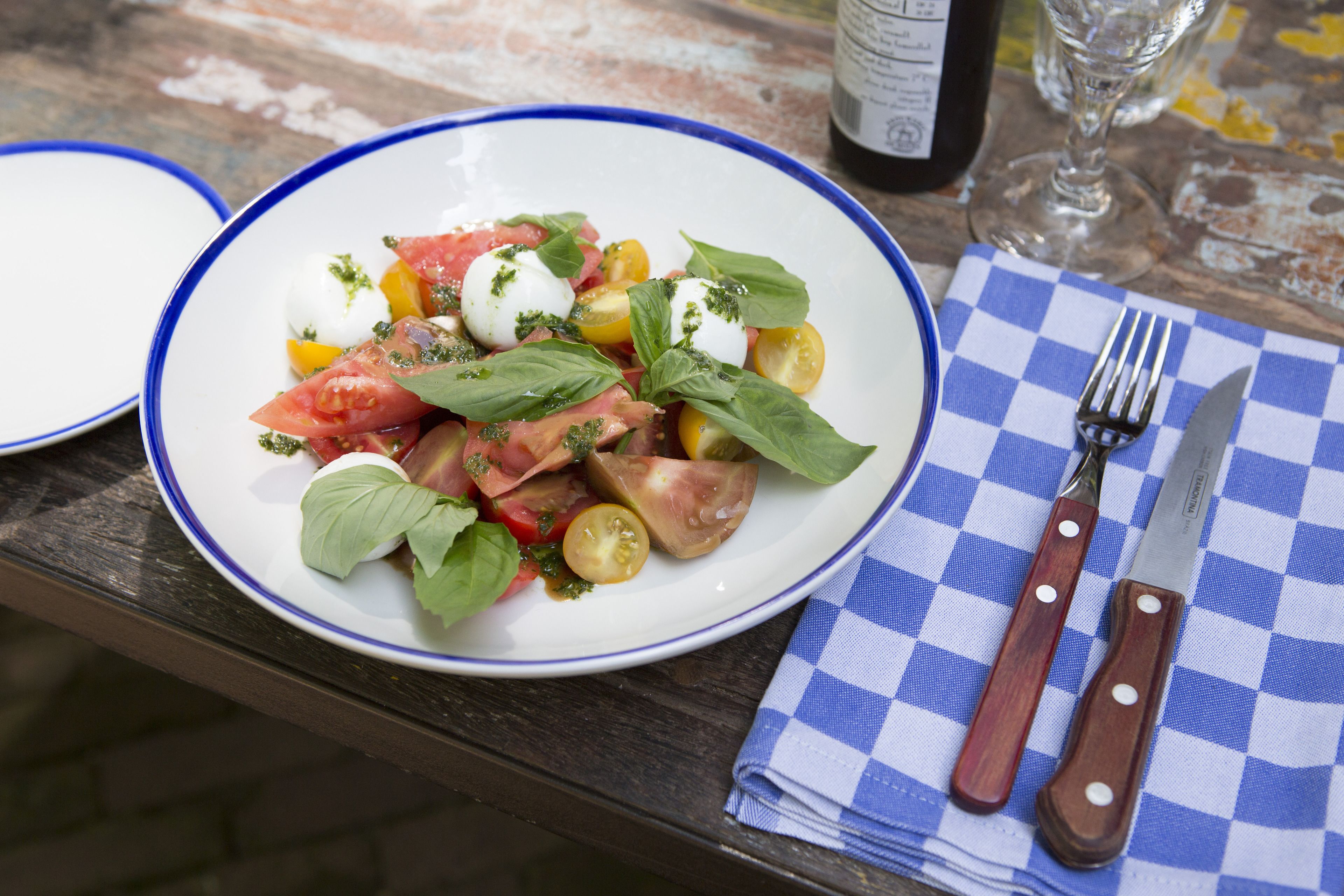 A plate of Caprese salad with tomatoes, mozzarella, and basil, accompanied by a fork and knife on a checkered napkin.