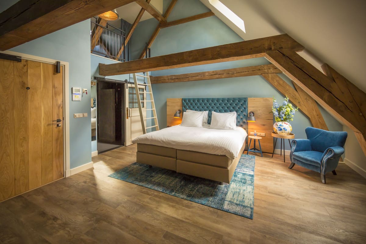 A bedroom with a large bed, wooden beams, a small chair, and a ladder leading to a loft.