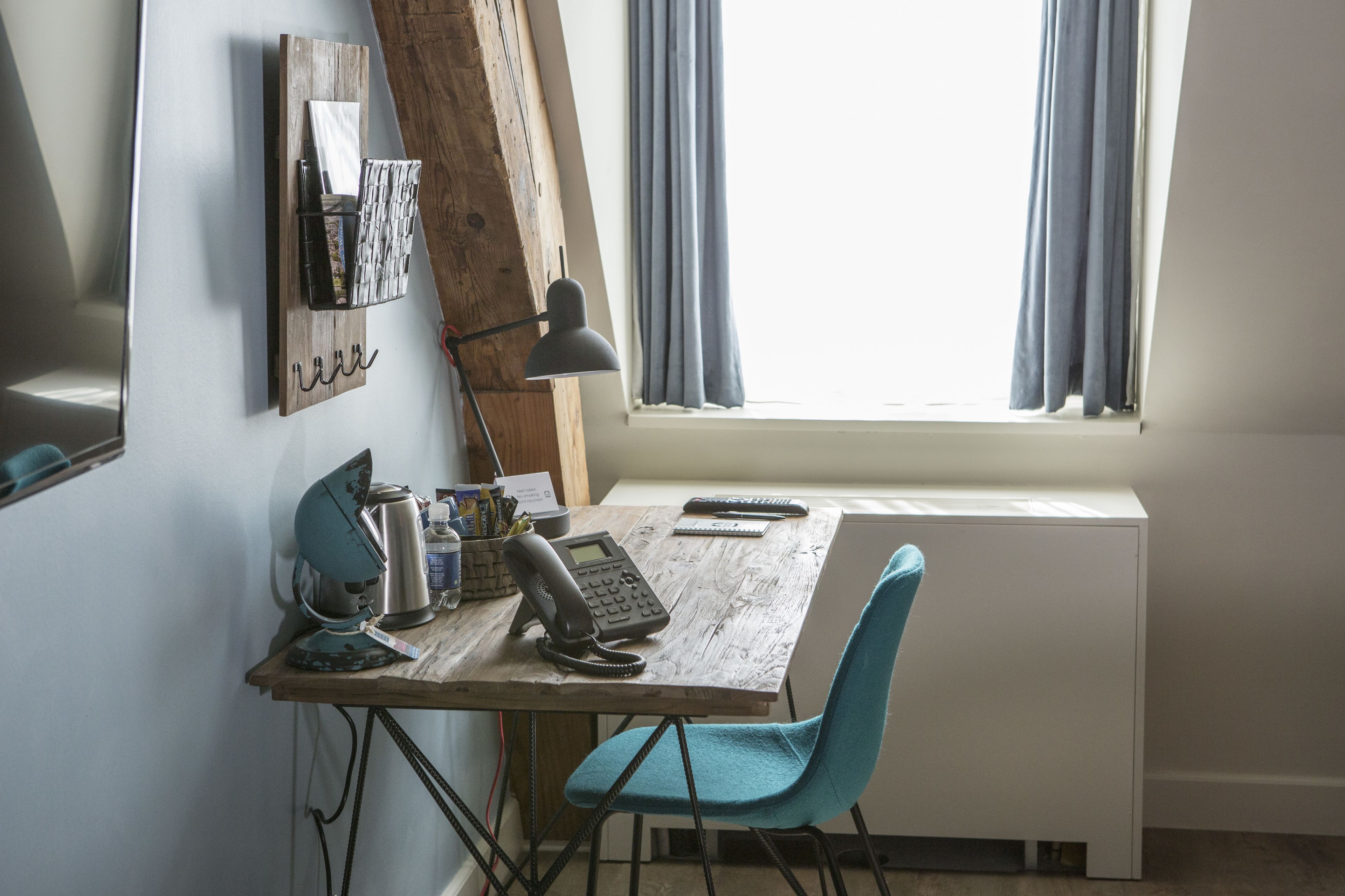 Un petit bureau rustique, avec un bureau en bois, une chaise bleue, un téléphone, une lampe et une fenêtre avec des rideaux.