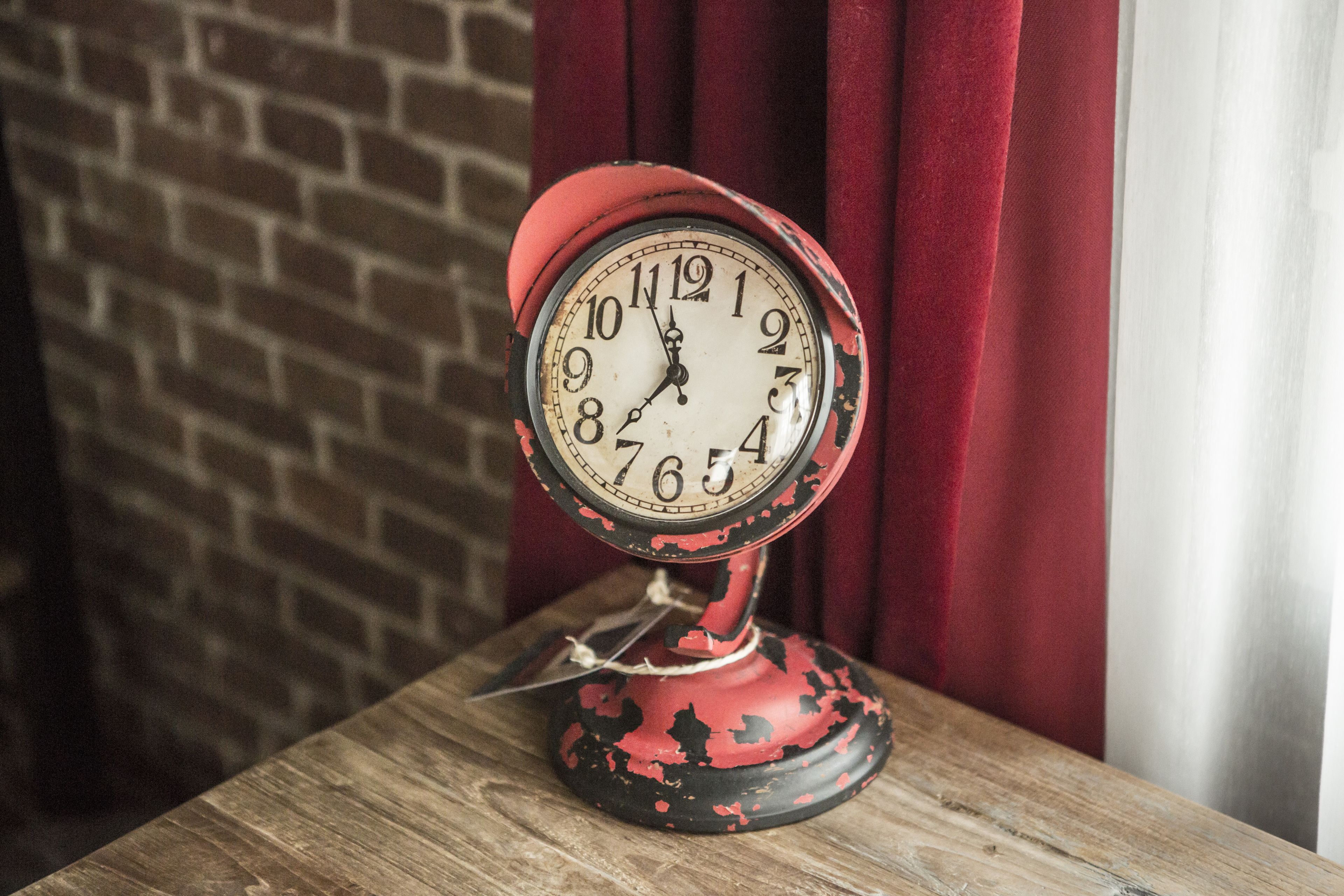 Un reloj antiguo rojo con esfera blanca y números negros, colocado sobre una mesa de madera frente a una pared de ladrillo y cortinas rojas.