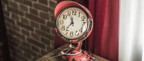 Un reloj antiguo rojo con esfera blanca y números negros, colocado sobre una mesa de madera frente a una pared de ladrillo y cortinas rojas.