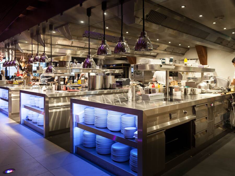 A modern kitchen with stainless steel appliances, overhead purple pendant lighting, and neatly stacked white plates on display.