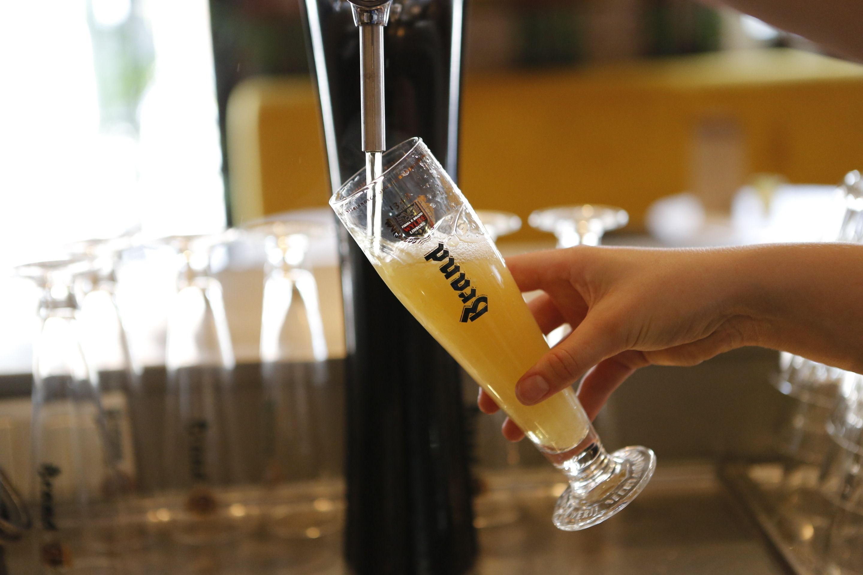 A hand holding a glass with a beverage, with a tap and glasses in the background.
