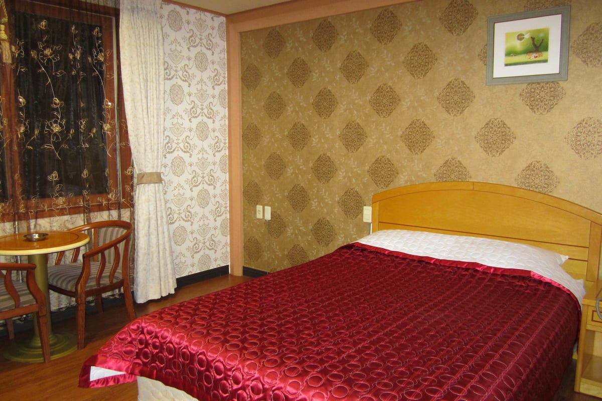 Habitació d'hotel amb un capçal de llit de fusta, un llençol vermell amb disseny, una cadira de fusta i una taula.
