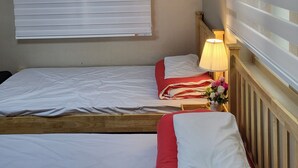 Habitación con dos camas, cada una con ropa de cama blanca y detalles rojos, una mesita de noche con lámpara y una ventana con persianas.