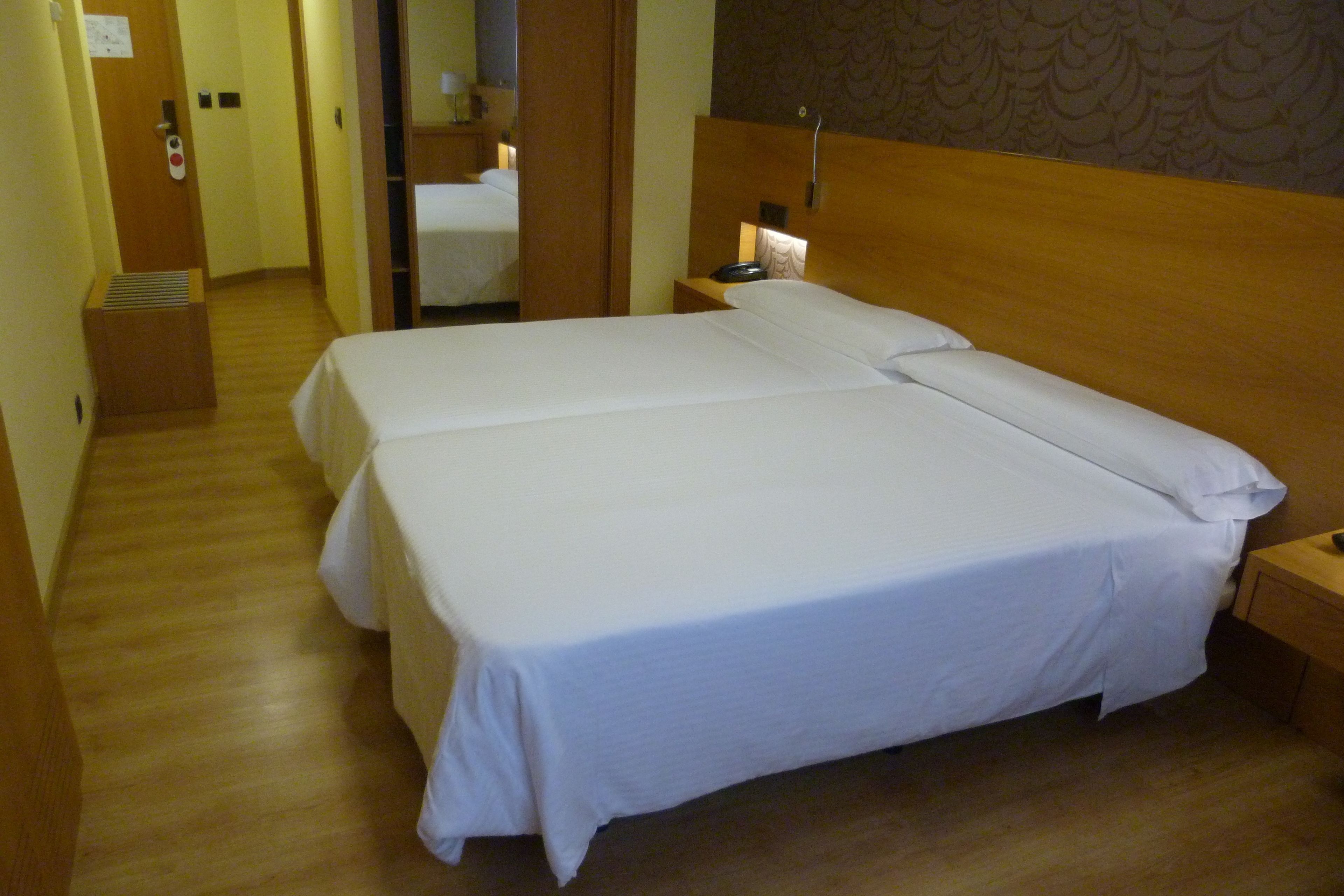 Habitación de hotel con dos camas, piso de madera, una mesita de noche con lámpara y una puerta con botón de emergencia rojo.