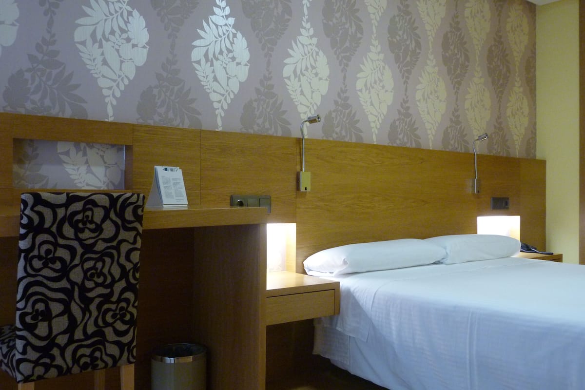A hotel room with a bed, a nightstand, a lamp, a chair, and a trash can.