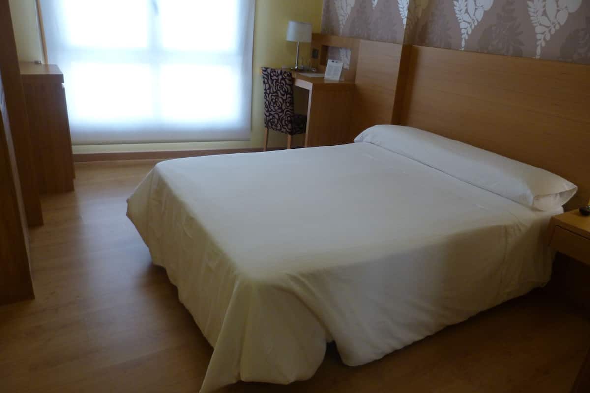 A neatly made bed in a hotel room with a wooden headboard, a bedside table with a lamp, and a chair with a patterned cushion.