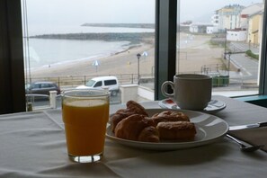 A breakfast table with croissants and a glass of orange juice, overlooking a beach and a pier.
