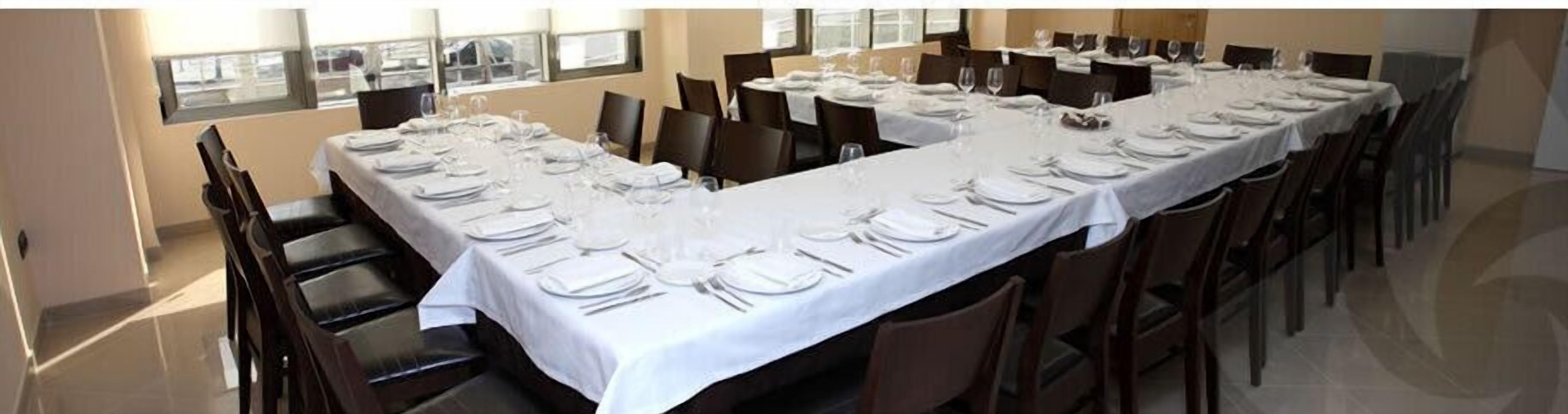 Una larga mesa de comedor con manteles blancos, varias sillas y vistas al exterior a través de amplios ventanales.