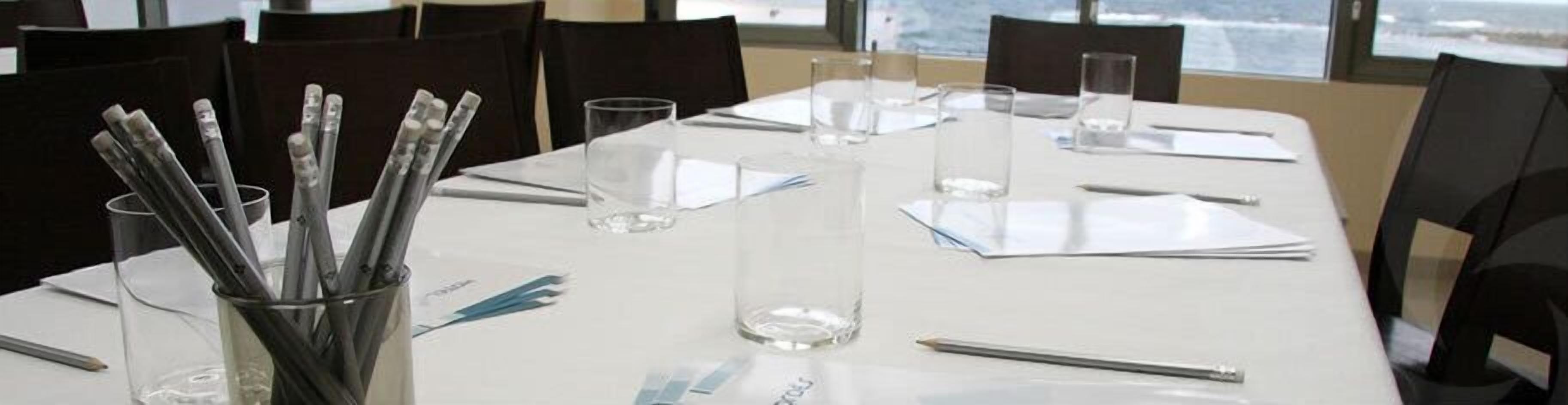 A conference table set with clear glasses, pens, and papers, in a room with large windows and black chairs.