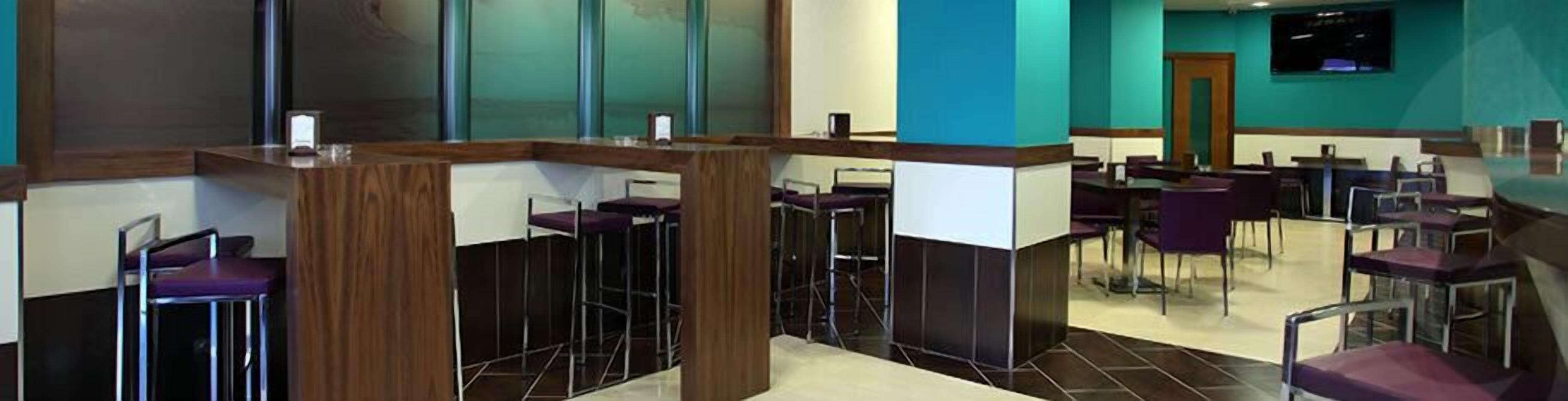Un moderno vestíbulo de hotel con techos altos, un mostrador de recepción central y varias zonas de estar con taburetes y mesas.