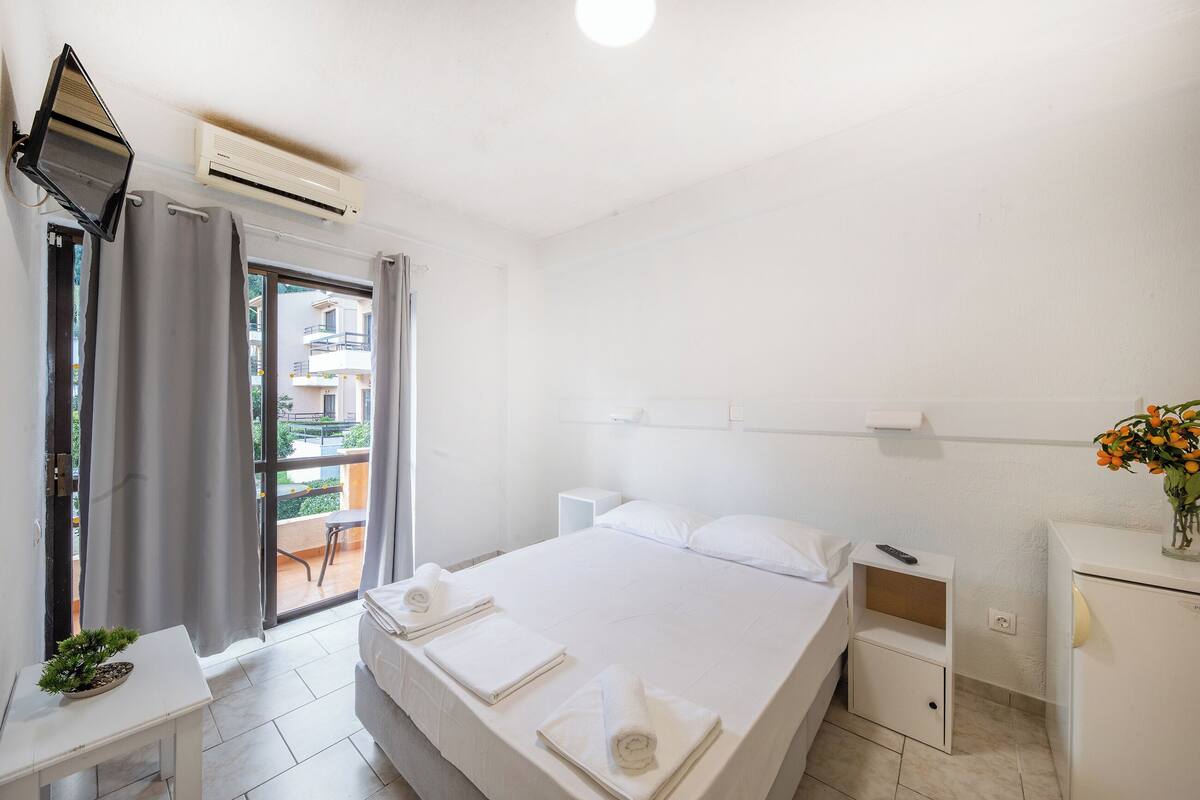 A hotel room with a bed, a small table with a plant, a nightstand, a TV, and a balcony with curtains.