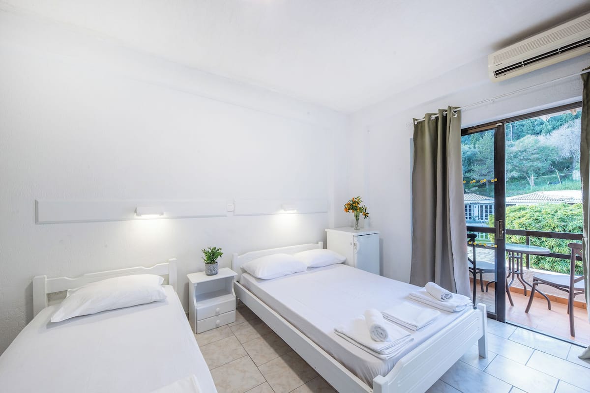A hotel room with two beds, a small table with a plant, a cabinet, and a balcony with a view of greenery.