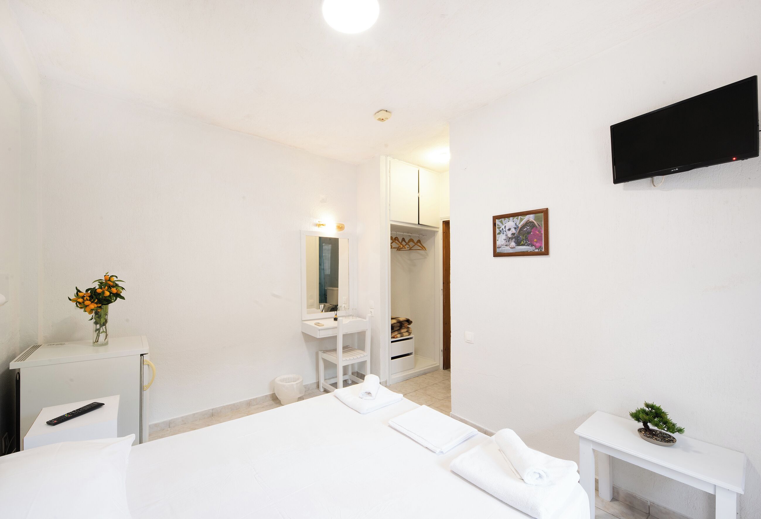 A hotel room with a bed, a bedside table with a remote control, a small desk with a vase of flowers, a white cabinet, a mirror, a television mounted on the wall, and a framed picture.