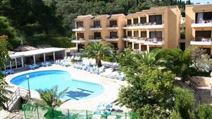 Un hotel con piscina circondata da poltrone a sdraio e palme, con sullo sfondo colline e vegetazione.