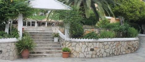 A stone staircase leading to a covered area with a white railing, surrounded by potted plants and palm trees.