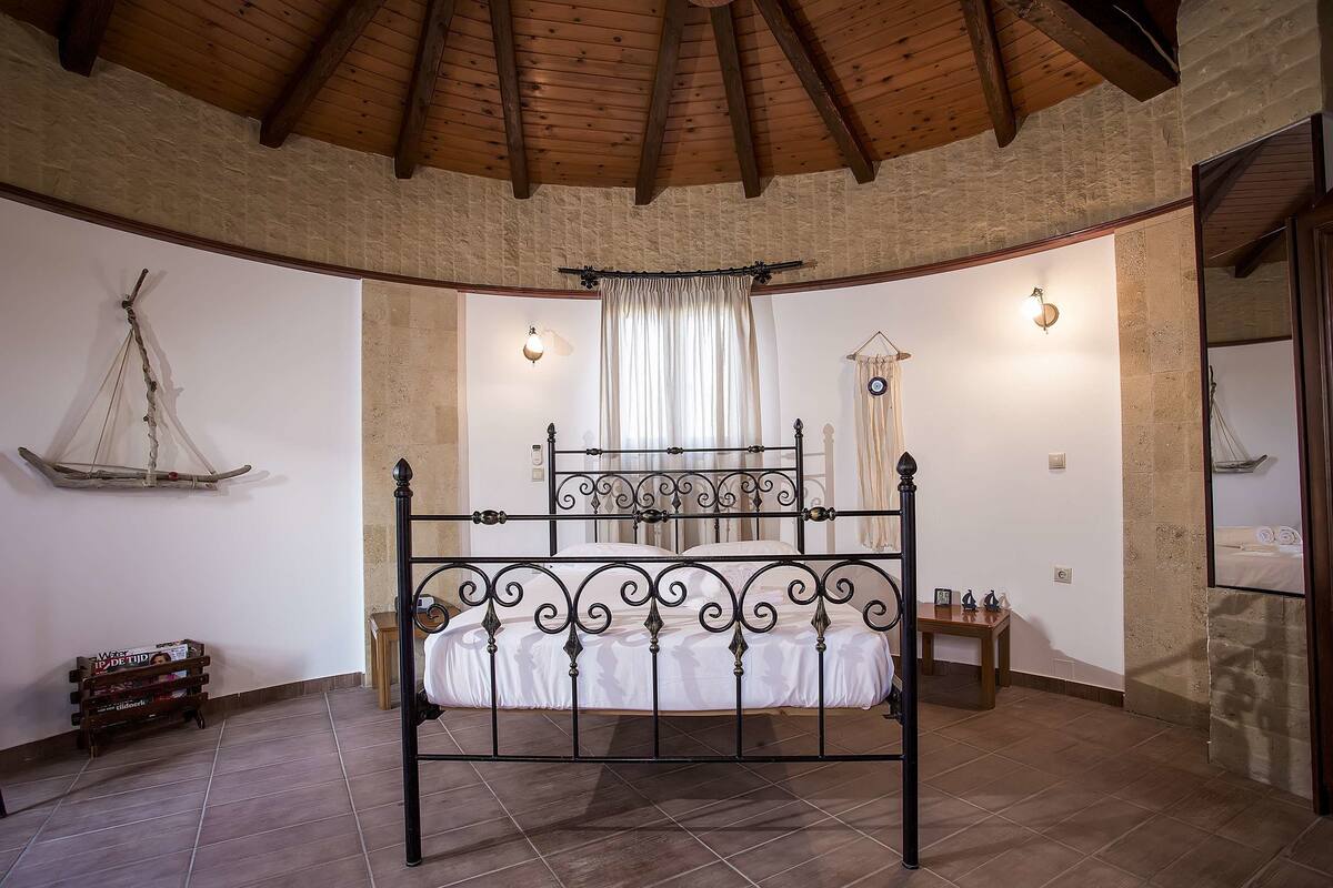 Quarto com teto curvo, cama com estrutura metálica, mesa de cabeceira pequena e prateleira fixada na parede.