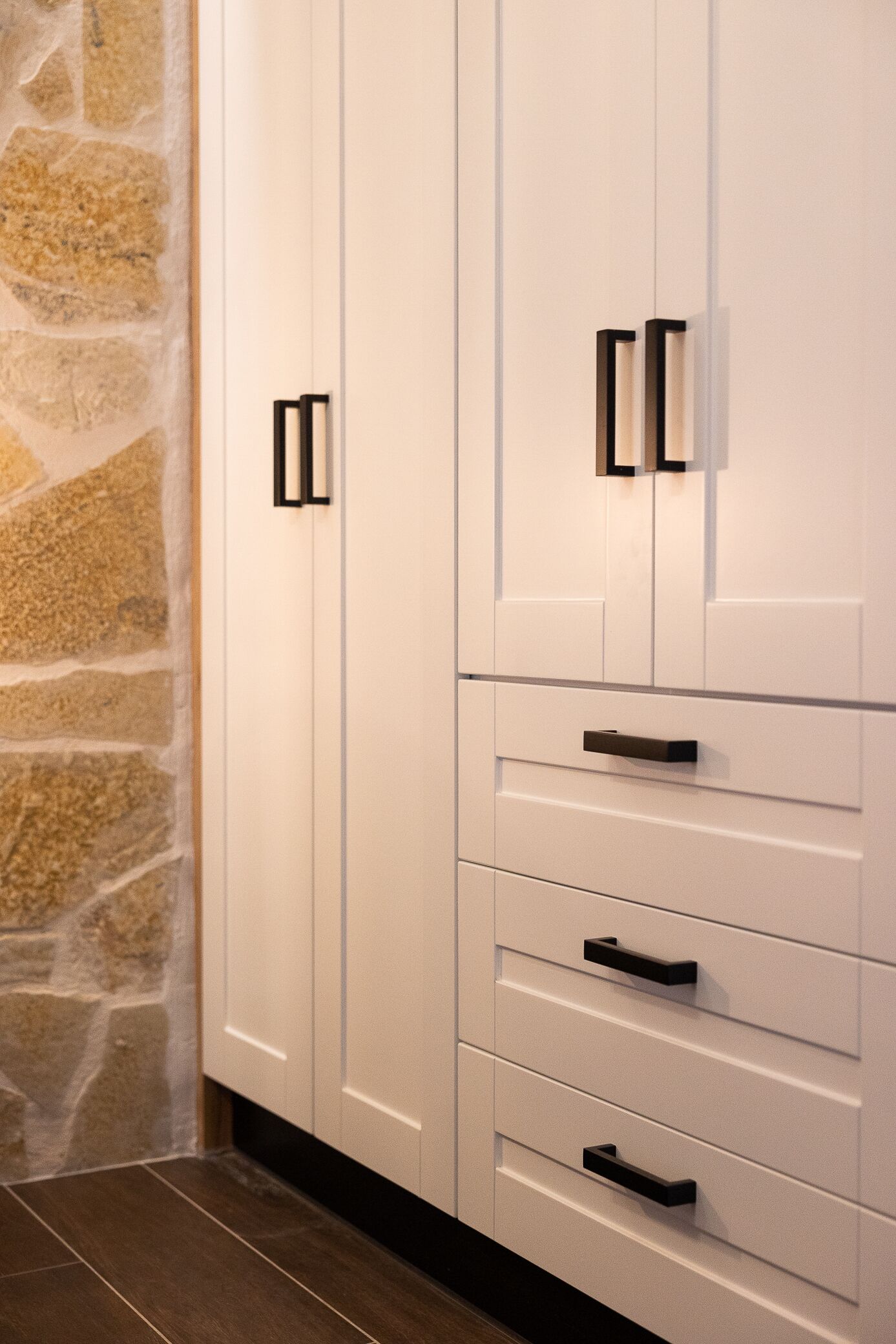 A set of white cabinets with black handles against a stone wall.