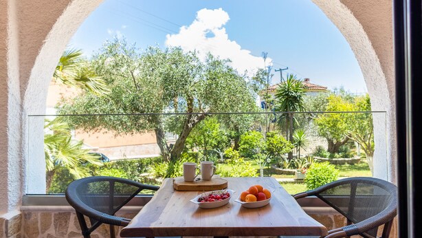 A balcony with a wooden table, two chairs, a bowl of fruit, and a glass railing overlooking a garden with trees and a building in the background. - Mathraki Resort (Corfu)