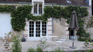 A building with a white facade, a large window, and a door with a metal gate. There are outdoor tables and chairs, and a potted plant in the foreground. - Le Moulin du Bas Pesé (Mont-pres-Chambord)