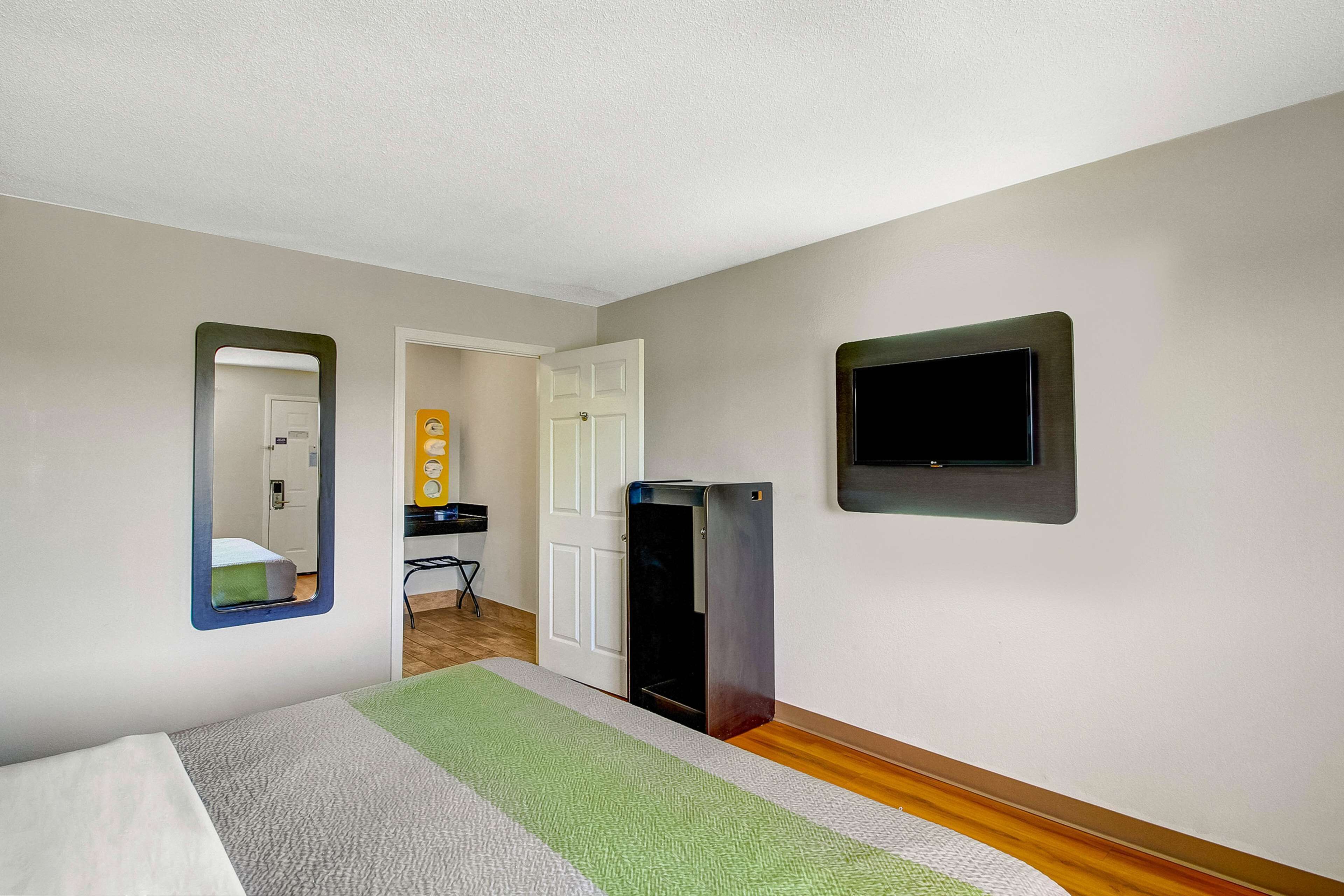 A hotel room with a bed, a small desk, a mirror, a television, and a door leading to another room.