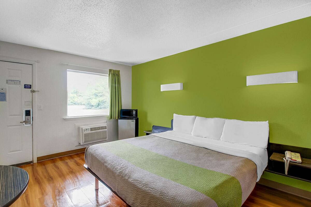 Een hotelkamer met een bed, bureau, stoel, raam met gordijnen, magnetron en thermostaat.