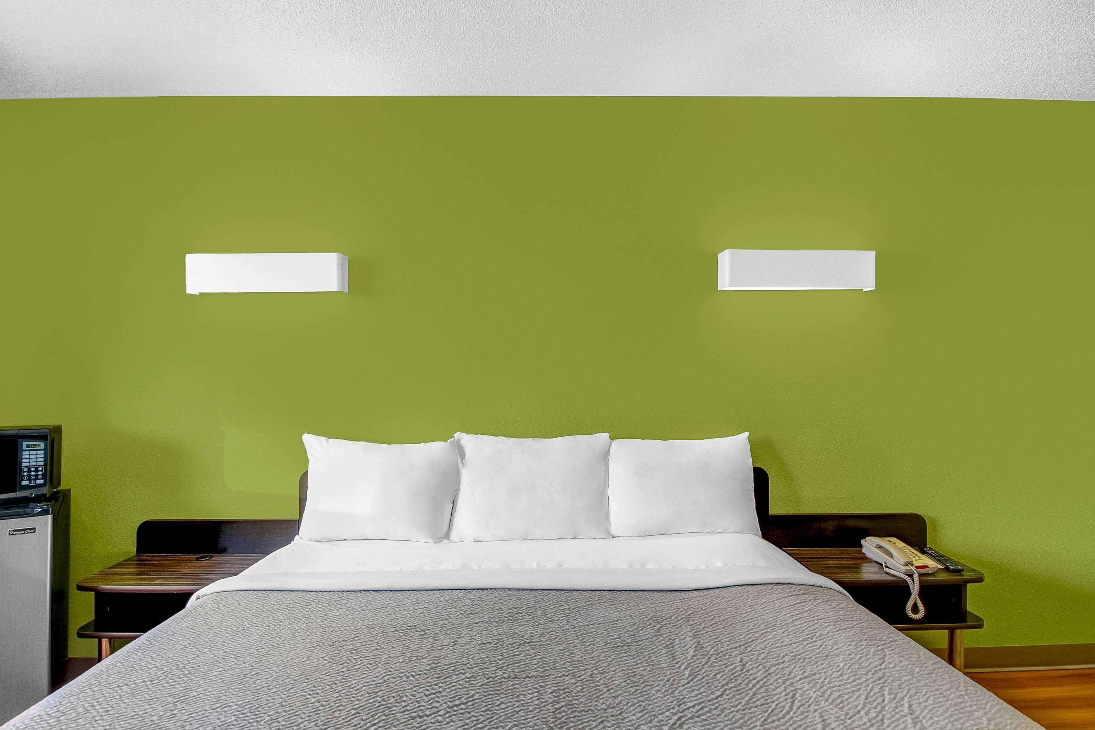 A hotel room with a bed, a bedside table with a phone, and a small desk with a computer.