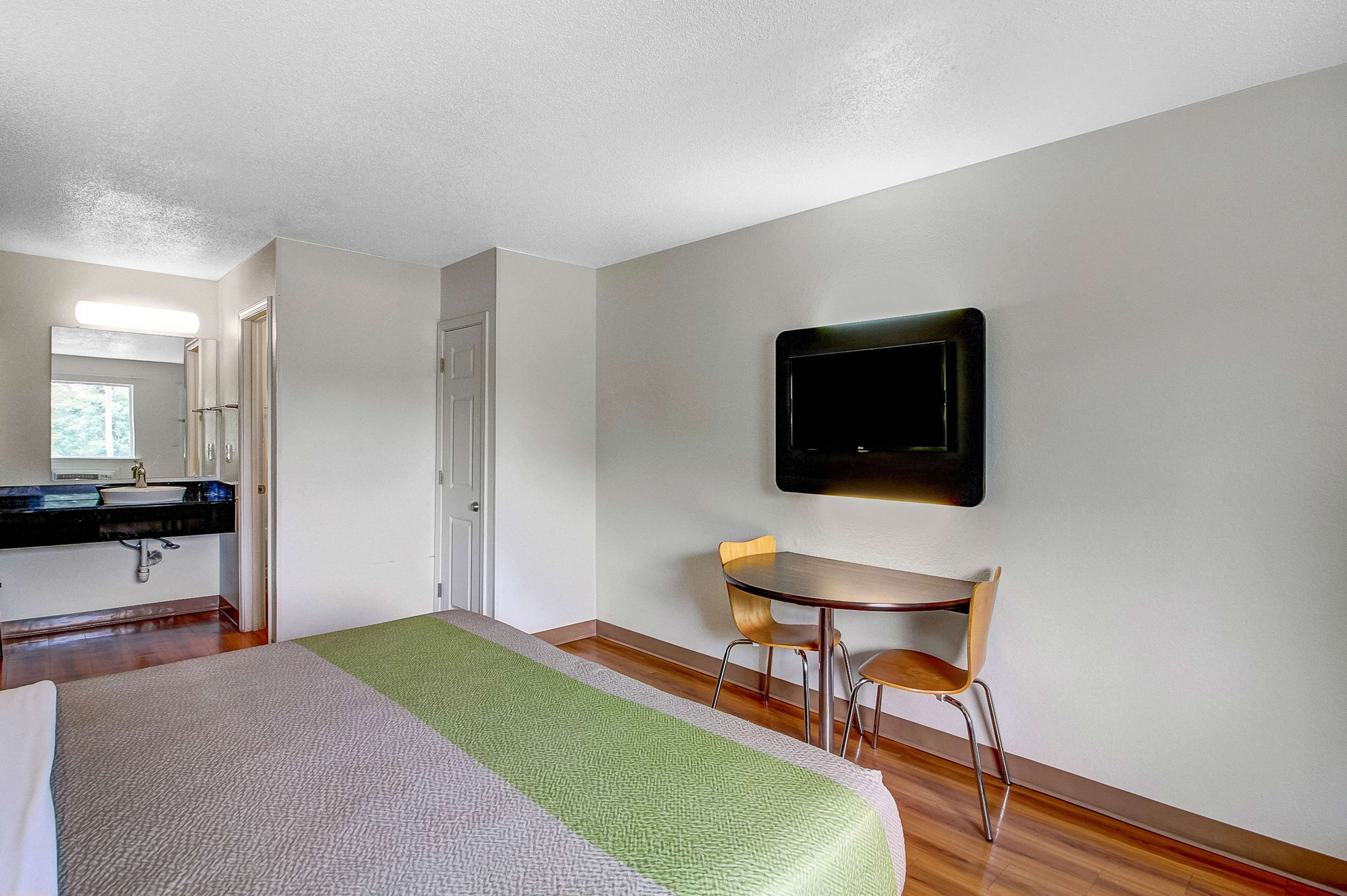 Habitación de hotel con cama, escritorio, silla, televisor montado en la pared y baño con lavamanos y espejo.